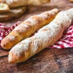 Frische Baguettes mit Mehlstaub auf Holzoberfläche neben kariertem Tuch – einfaches Baguette Rezept.