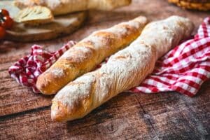Zwei knusprige Baguettes mit Mehlstaub auf Holzuntergrund und rot-weiß kariertem Tuch zum Baguette Rezept.