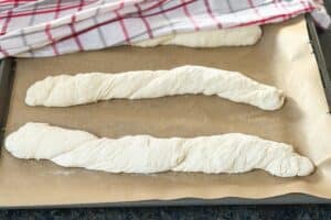 Drei gedrehte Baguette-Stränge aus Hefeteig vor dem Backen auf Backpapier.