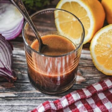 Balsamico-Dressing in einem Glas mit Zitrone und Zwiebel auf Holztisch neben rot-weiß kariertem Tuch.