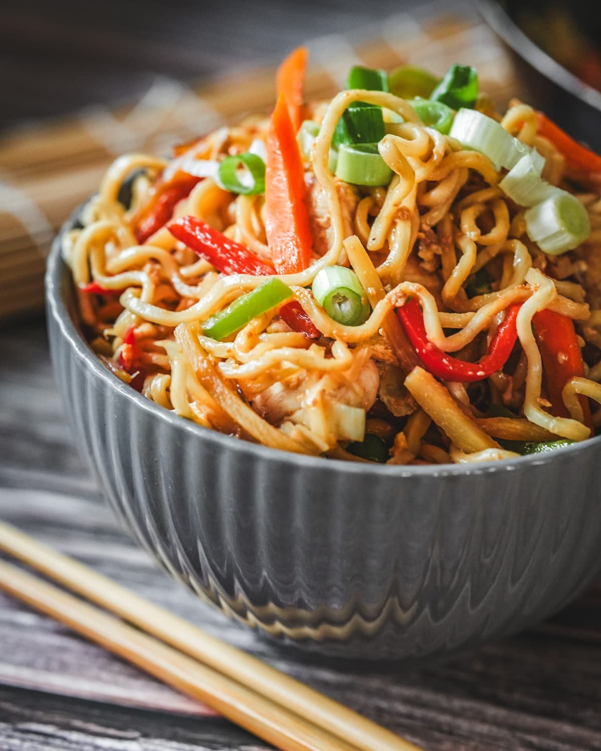 Bami Goreng mit Hähnchen in grauer Schüssel, garniert mit Frühlingszwiebeln und Paprika, serviert mit Stäbchen.