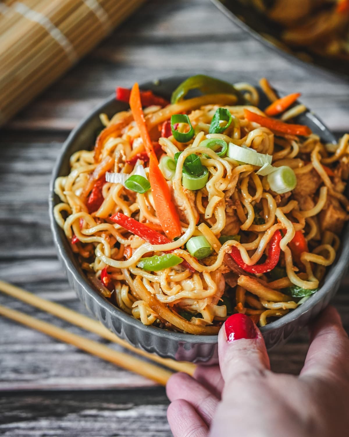 Gebratene Bami Goreng mit Hähnchen, Paprika und Frühlingszwiebeln in einer grauen Schale mit Stäbchen.