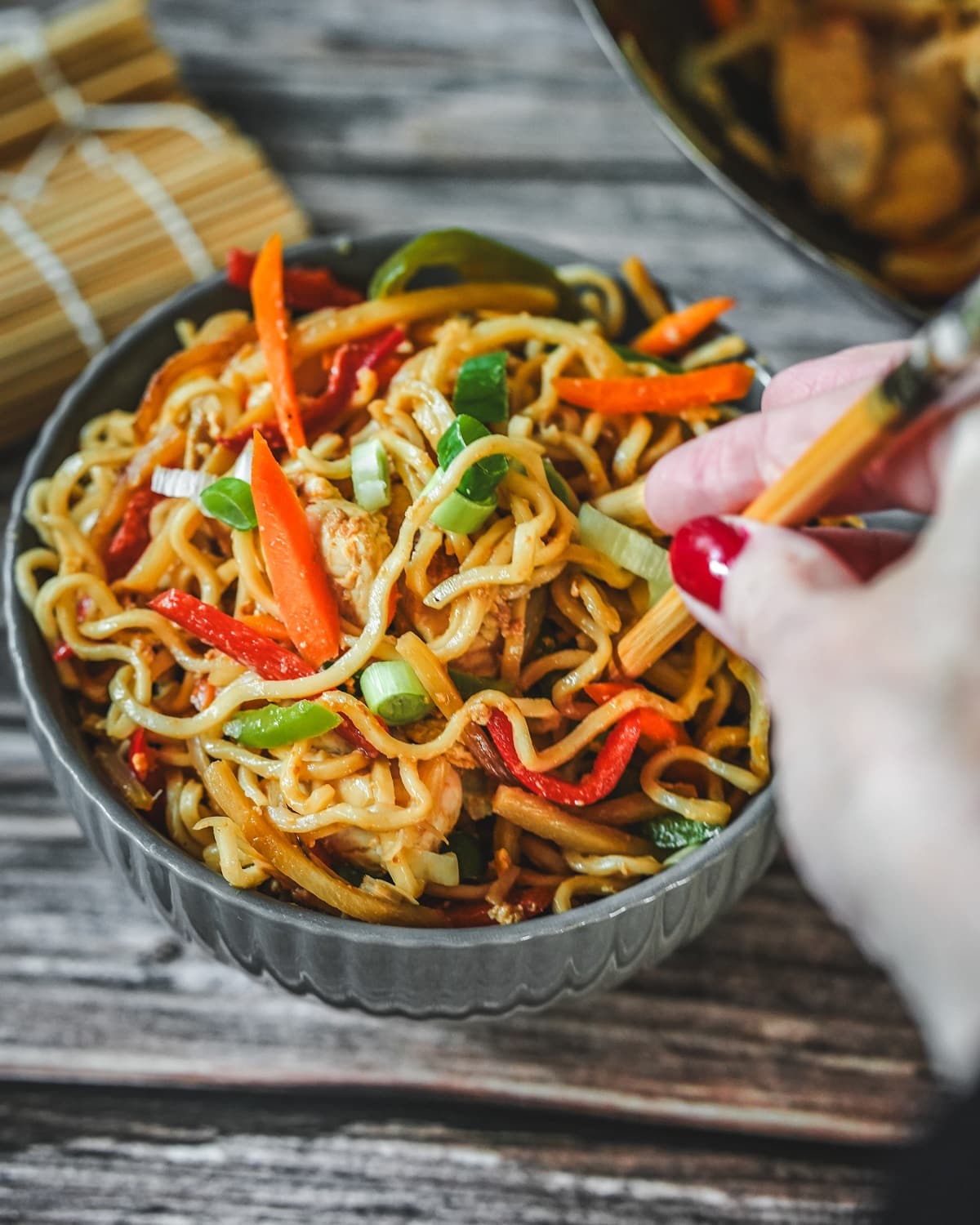 Gebratene Bami Goreng mit Hähnchen, Paprika und Frühlingszwiebeln in grauer Schale mit Stäbchen.
