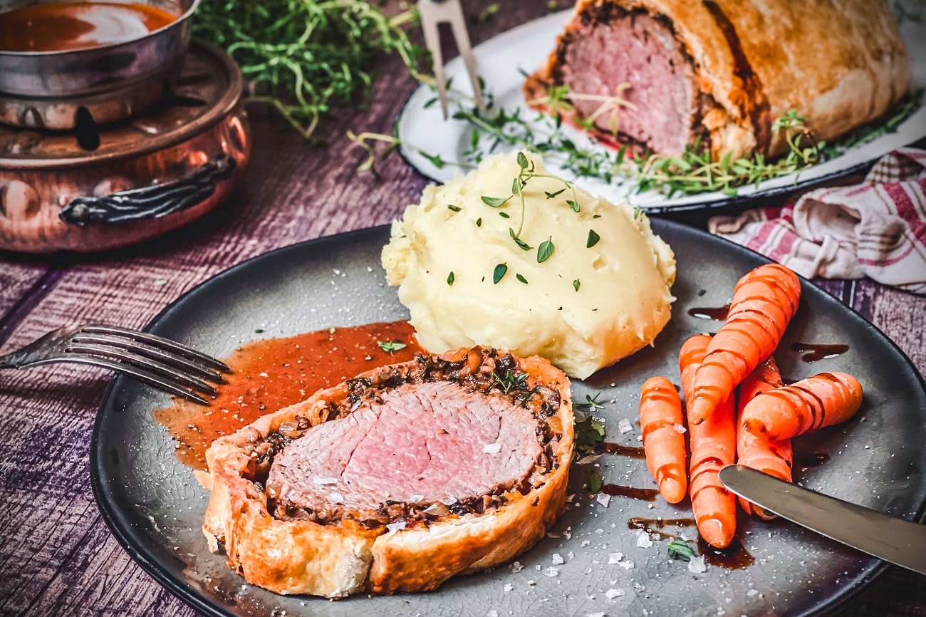 Filet Wellington mit rosa Fleischkern, Kartoffelpüree und Karotten auf schwarzem Teller serviert.