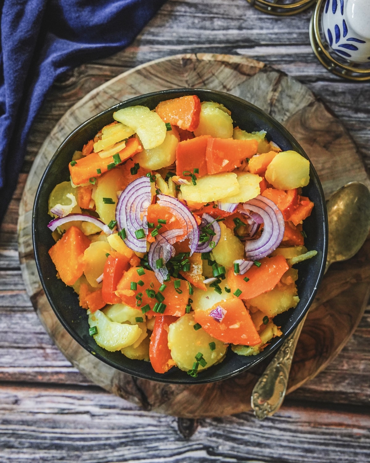 Hokkaido-Kürbis-Kartoffel-Salat mit roten Zwiebeln und Schnittlauch in blauer Schüssel auf Holzbrett.