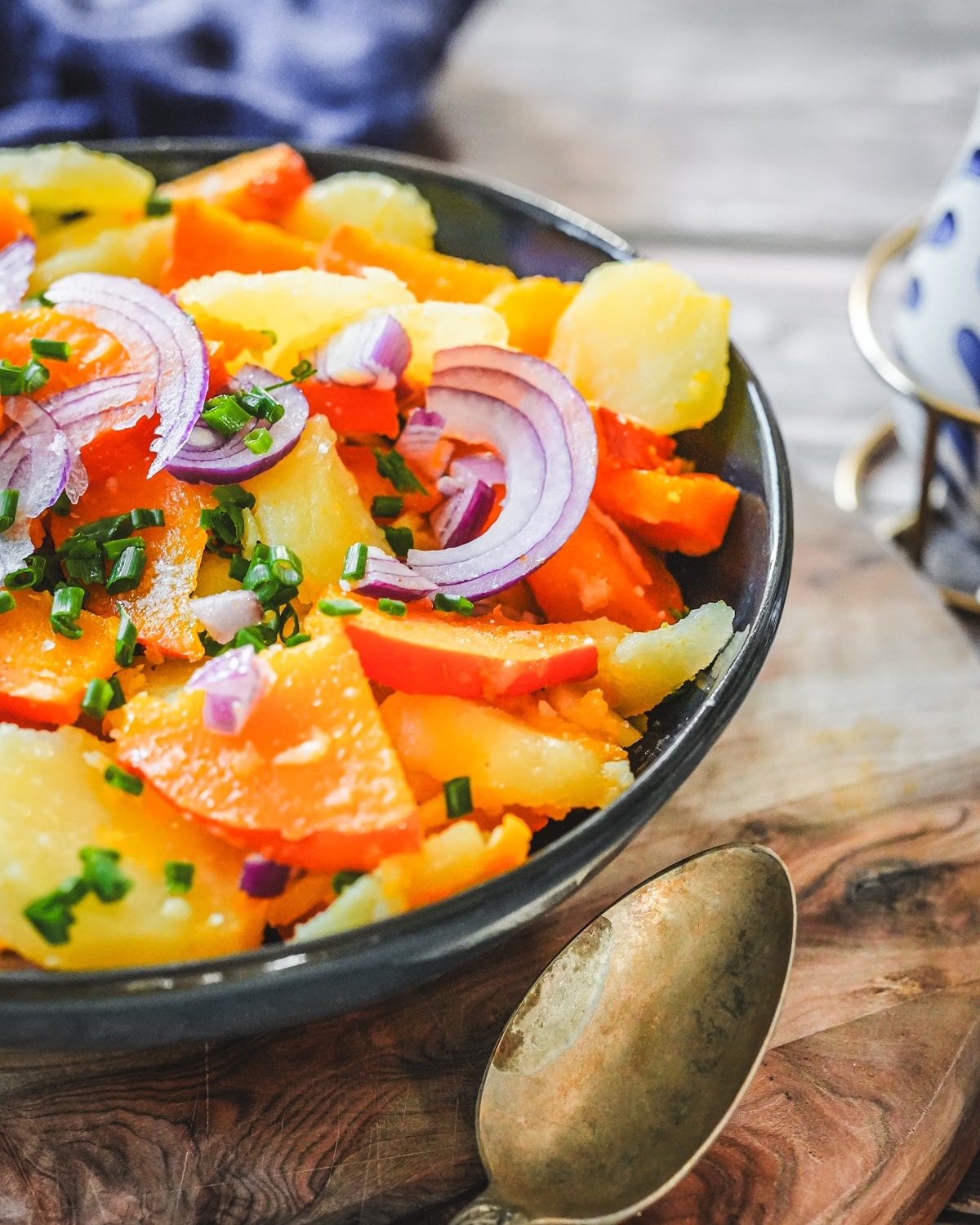 Kürbis-Kartoffel-Salat mit roten Zwiebeln und Schnittlauch in blauer Schale auf Holztisch.