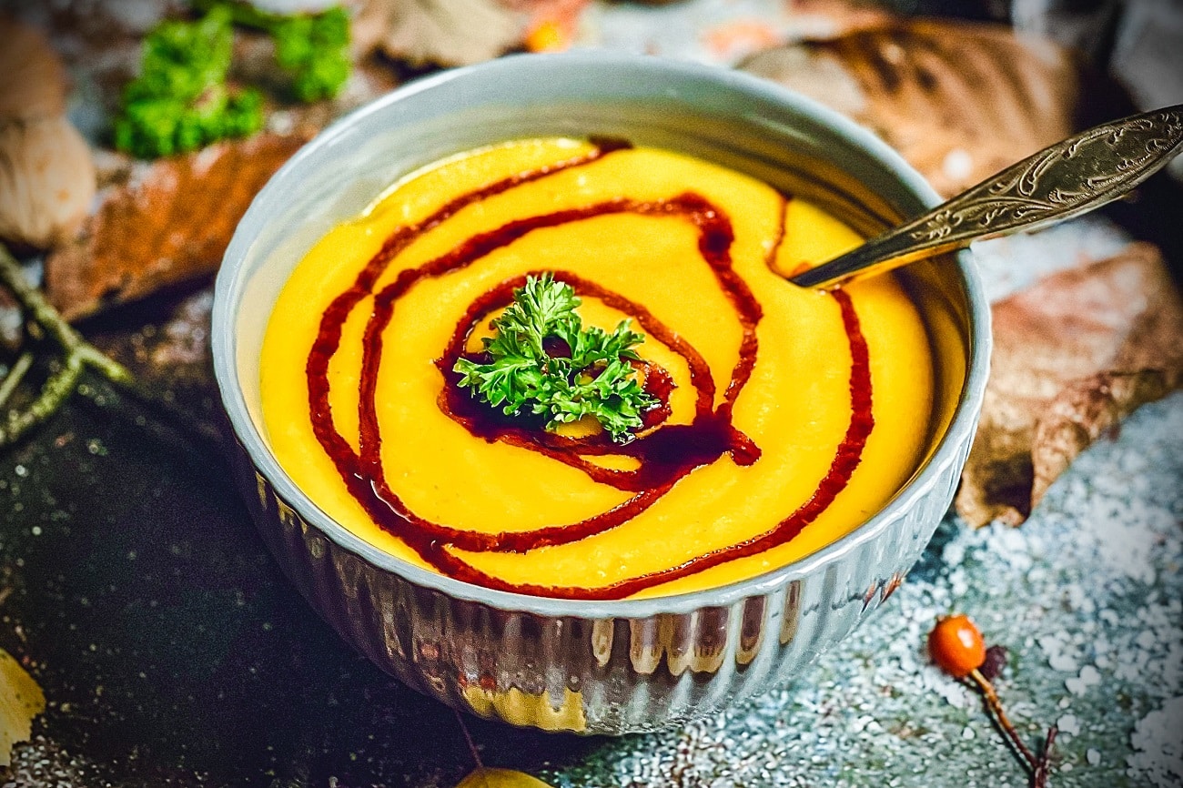 Cremige Kürbissuppe in grauer Schüssel mit rotem Öl-Swirl und frischer Petersilie garniert.