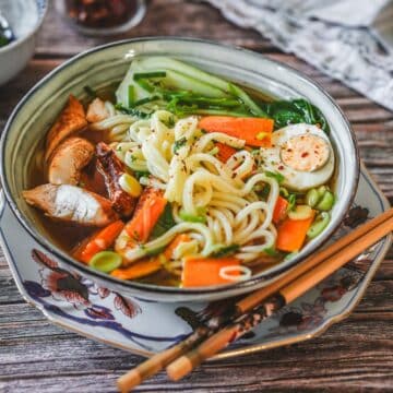 Schüssel mit Ramen Suppe bestehend aus Nudeln, Hähnchen, Gemüse und Brühe, garniert mit Kräutern und Chili.