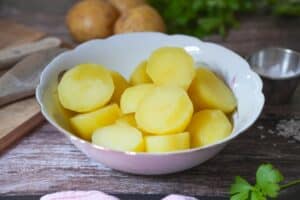 Gekochte Salzkartoffeln in einer rosa Schüssel, angerichtet mit frischer Petersilie und Rohkartoffeln im Hintergrund.