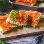 Spanakopita-Stücke mit goldbraunem Filo-Teig, Sesam und frischer Petersilie auf Holzbrett serviert.