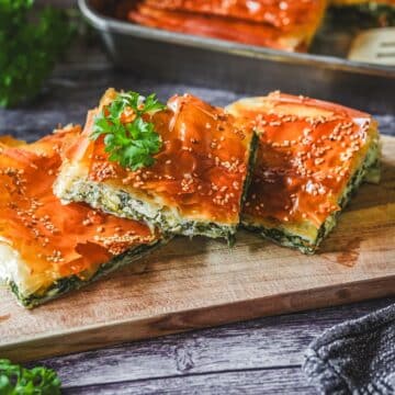 Spanakopita-Stücke mit goldbraunem Filo-Teig, Sesam und frischer Petersilie auf Holzbrett serviert.