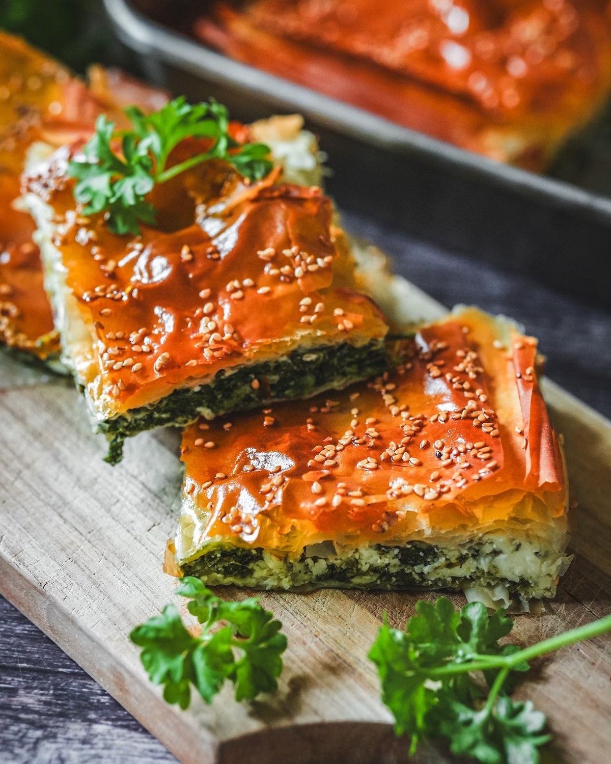 Spanakopita-Stücke mit goldbraunem Blätterteig, Sesam und frischer Petersilie auf Holzbrett serviert.