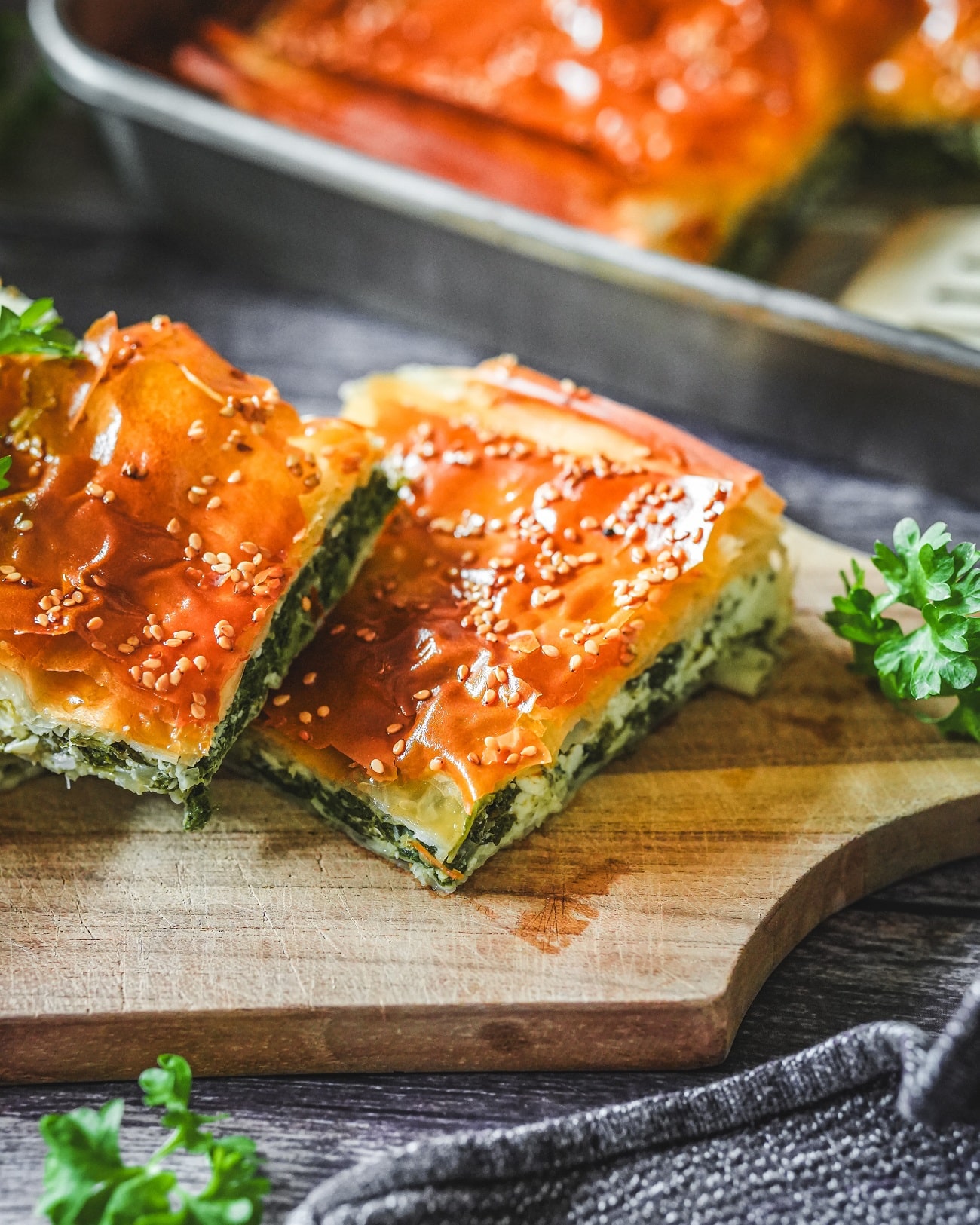 Spanakopita-Stücke mit goldbraunem Blätterteig, Spinat-Feta-Füllung und Sesam auf Holzbrett serviert.