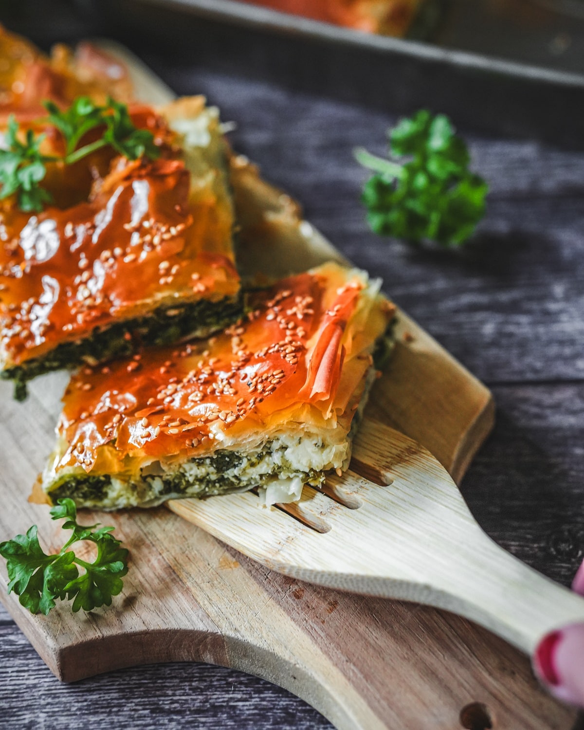 Spanakopita-Scheiben mit knusprigem Blätterteig, Spinat-Feta-Füllung und Sesam auf Holzbrett garniert.