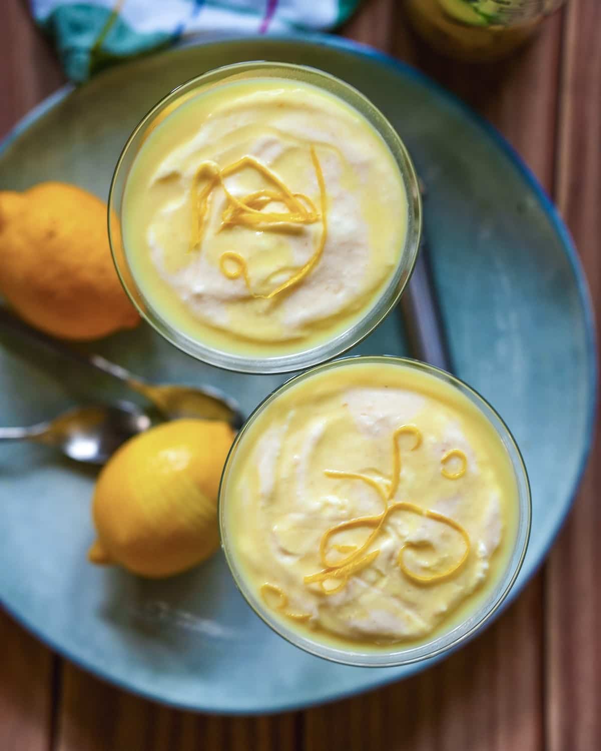 Zitronencreme-Dessert mit Zitronenschale-Garnierung in Gläsern auf blauem Teller mit frischen Zitronen.