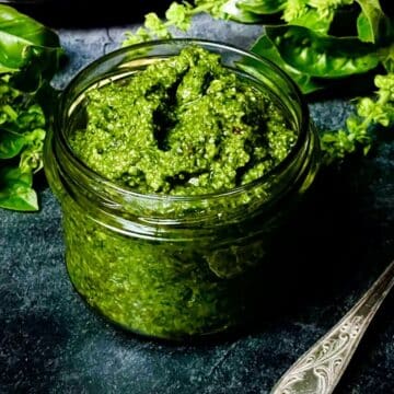 Basilikum Pesto in einem Glas mit frischen Basilikumblättern und Besteck auf dunklem Untergrund.
