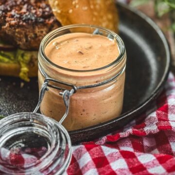 Hausgemachte Grillsoße in Gläsern neben gegrilltem Burger auf Holzuntergrund mit Kräutern und kariertem Tuch.