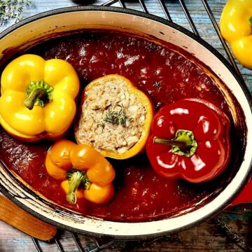 Gefüllte Paprika in Tomatensoße köcheln in einem roten Bräter auf Holzuntergrund.
