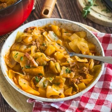 Kartoffelpfanne mit Hackfleisch in weißer Schüssel auf Holztisch mit rotem KochTopf im Hintergrund.