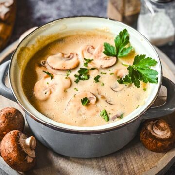 Cremige Pilzsuppe mit Champignons, Petersilie und Pfeffer in grauer Schüssel auf Holzbrett.