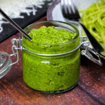 Frisches grünes Pesto im Glas mit Spoon, daneben Spaghetti und Küchenutensilien auf Holzoberfläche.