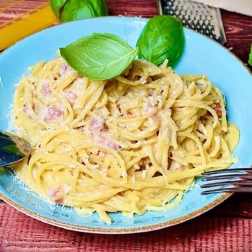 Spaghetti Carbonara mit Schinken auf blauem Teller, garniert mit frischem Basilikum auf Holztisch.